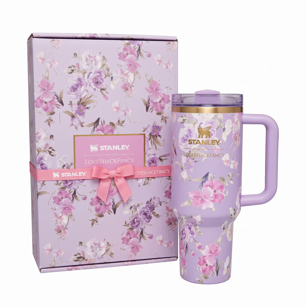 A lavender Stanley insulated tumbler with a pink floral print, shown next to its matching floral gift box with a pink ribbon and bow. The tumbler features a gold-toned band near the lid and the Stanley logo.