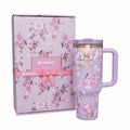 A lavender Stanley insulated tumbler with a pink floral print, shown next to its matching floral gift box with a pink ribbon and bow. The tumbler features a gold-toned band near the lid and the Stanley logo.