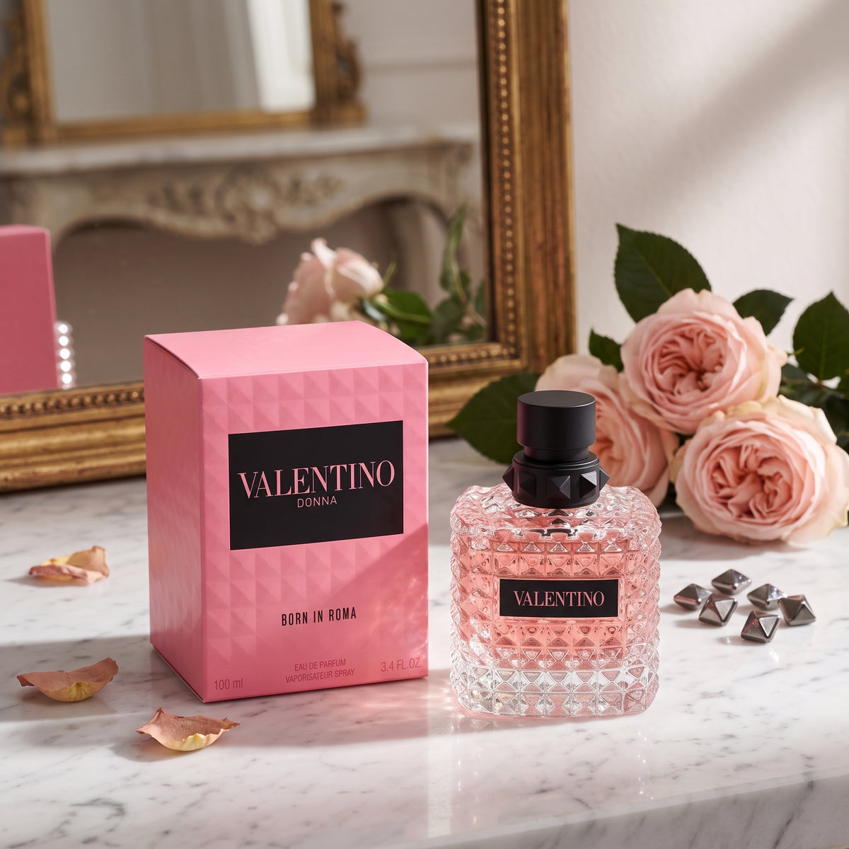 Valentino Donna Born in Roma Eau de Parfum bottle and its pink box, styled on a marble surface with pink roses and metallic studs.