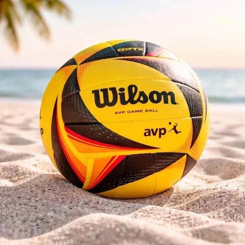 A Wilson AVP game ball volleyball sits on sandy beach ground with the ocean and palm leaves blurred in the background.
