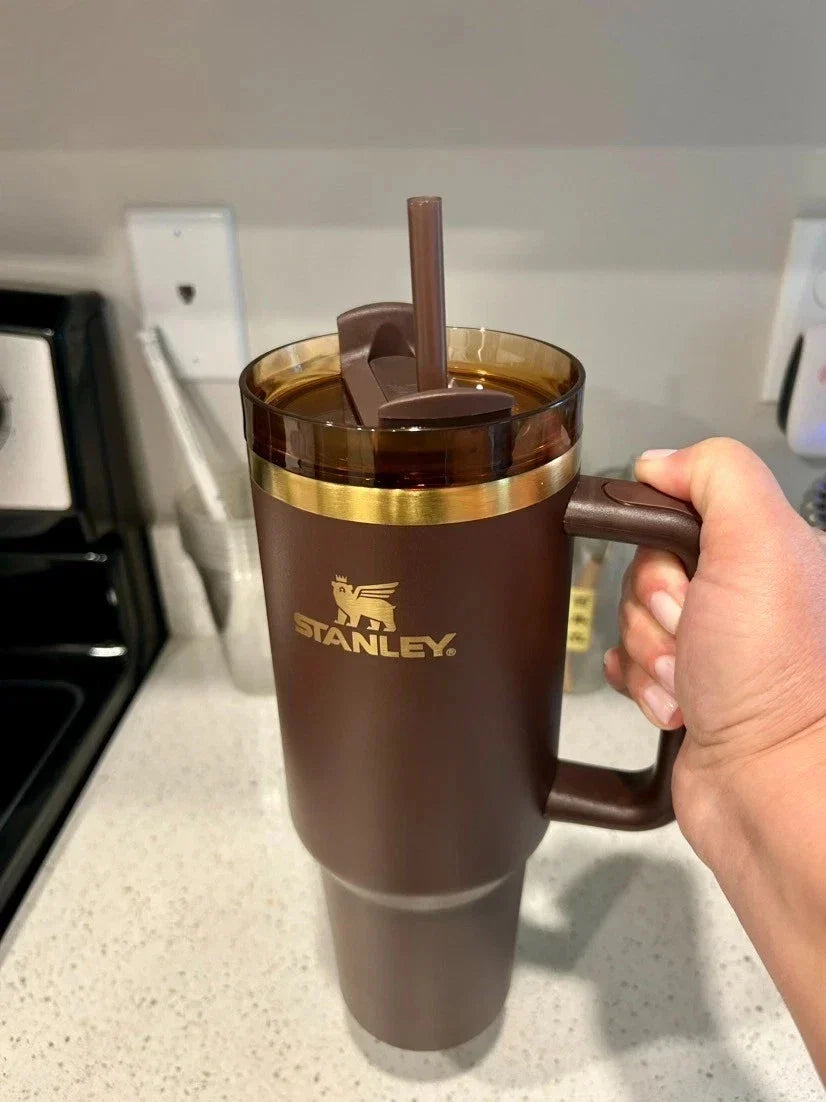A hand holding a brown Stanley travel tumbler with a handle, gold logo, and a matching brown straw lid, set against a kitchen countertop background.