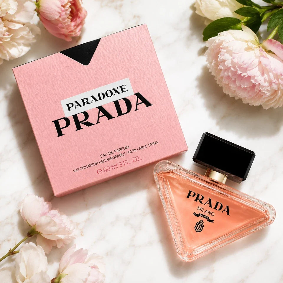 Prada Paradoxe Milano perfume bottle and its pink box, styled on a white marble surface with pink peony flowers.